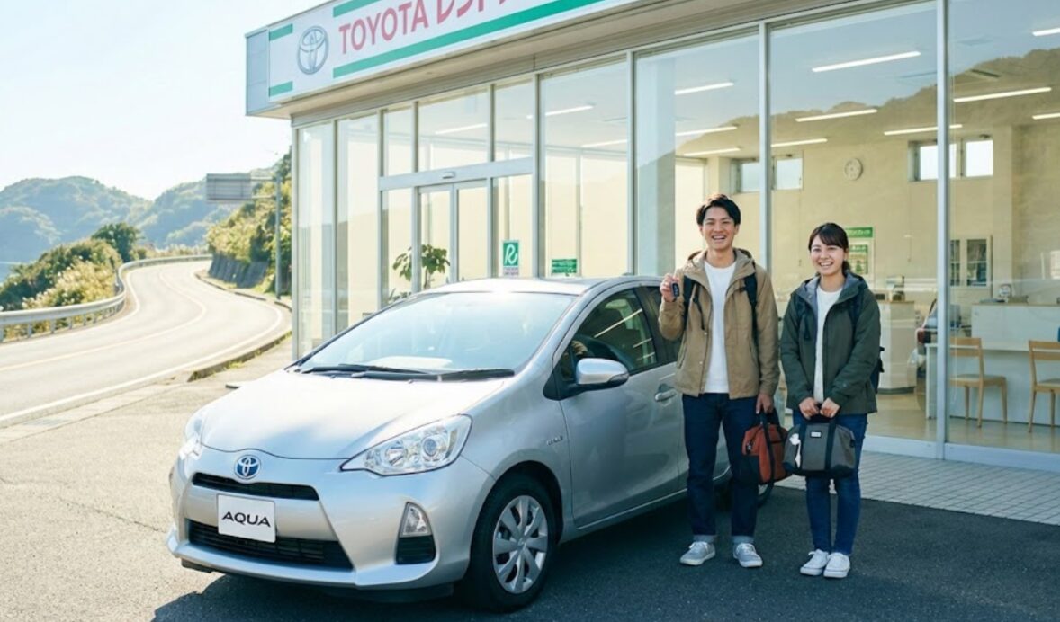 綺麗なトヨタレンタカーの営業所で、旅行に出発する前にレンタカー（ヤリス・アクア）の鍵を受け取る若い日本人カップル。